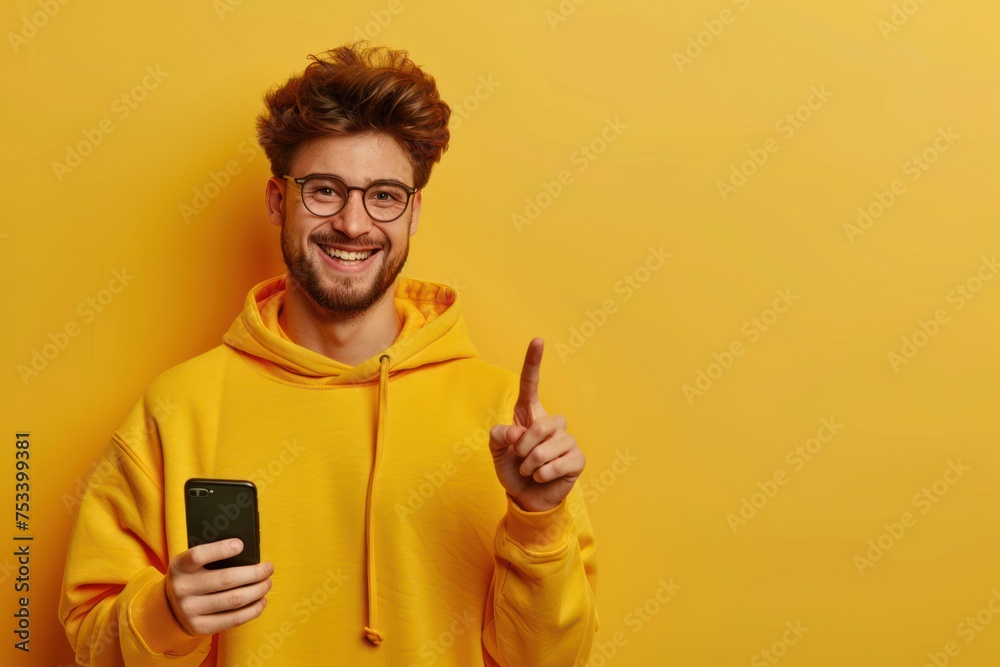 Portrait of young excited man pointing finger at smartphone. Cheerful guy in glasses and sweatshirt holding modern digital device and touching screen over yellow isolated studio background --ar 3:2