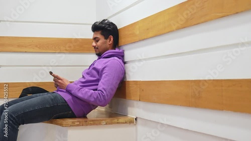 Guy reading on his phone while sitting in the corner of the room and leaning his back against the wall, static