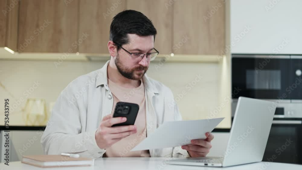 Competent caucasian male looking from document to modern smartphone during work process at personal space for freelancing at home. Bearded man successfully performing assigned tasks indoors. Competent caucasian male looking from document to modern smartphone during work process at personal space for freelancing at home. Bearded man successfully performing assigned tasks indoors.