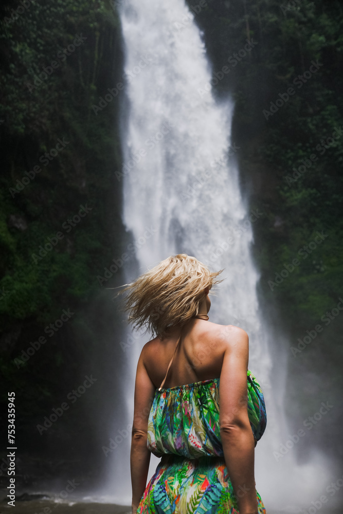 Luxurious blonde posing by a waterfall in a swimsuit, rear view.