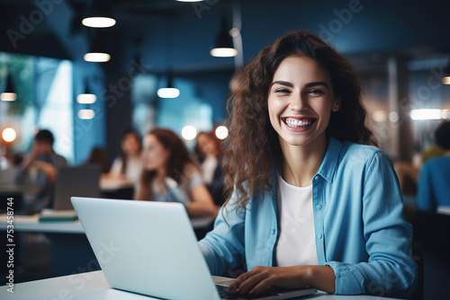 Young cheerful woman sitting in front of computer in casual clothes, smiling and working late at night at office. Looking at camera.