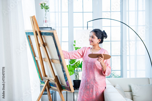 Cheerful Indian young artist girl painting picture at easel in living room photo