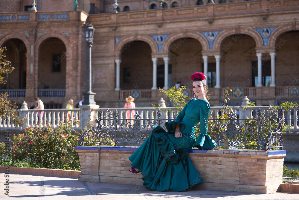 Young, pretty, blonde woman in typical green colored flamenco suit, posing sitting on a tiled and brick bench. Flamenco concept, typical Spanish, Seville, Andalusia.