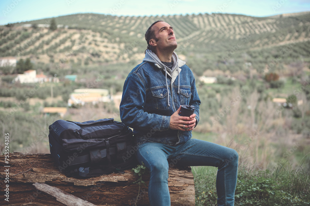 Happy young traveler man with backpack, sitting on a log in mountains, drinking coffee and breathing fresh air Happy young traveler man with backpack, sitting on a log in mountains, drinking coffee and breathing fresh air