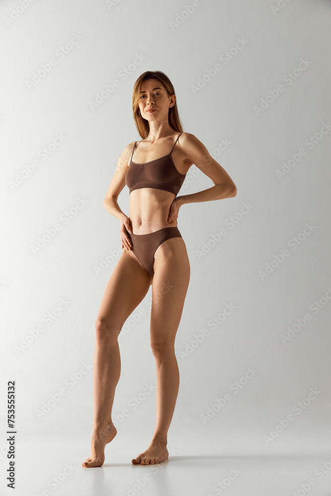 Full-length portrait of young woman in brown underwear, standing confidently with hands on hips against grey studio background. Concept of beauty treatments, dieting, female health, spa procedures. Ad