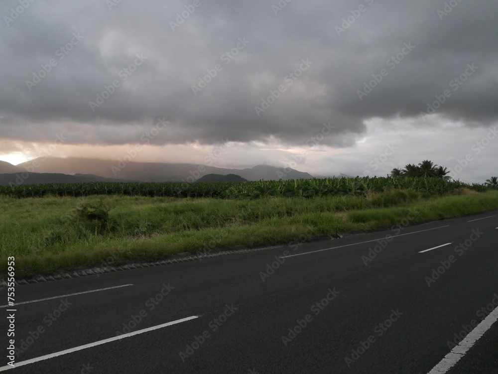 Naklejka premium empty highway and tropical field in basse terre, guadeloupe. Asphalt road with empty lanes and mountain background with storm clouds in dramatic sky