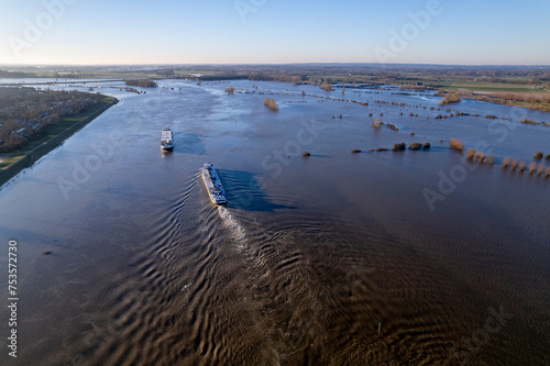 Inland shipping cargo ships passing the extended river IJssel during high water level with flood plains tripling the width of the fast flowing waterway. Dutch flat landscape aerial