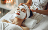 A woman getting a facial treatment at a spa salon. A beautiful woman lying on a white bed with a towel and enjoying a face mask at a beauty center. The hands of a professional beautician applying cosm