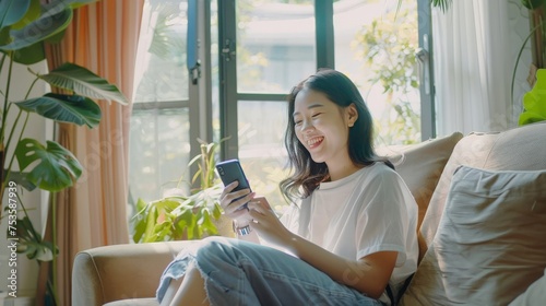 Dreamy asian girl spend time at home, holding smartphone and sitting on couch, smiling while looking at window
