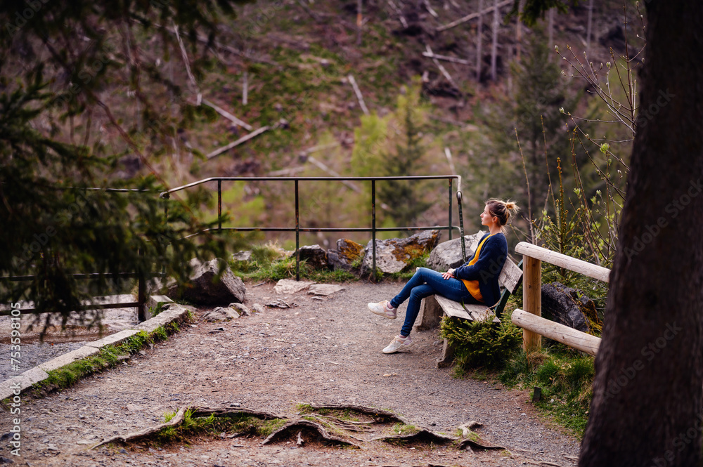 A woman takes a moment to rest on a wooden bench amidst a forested trail, absorbing the quietude and grandeur of the natural landscape