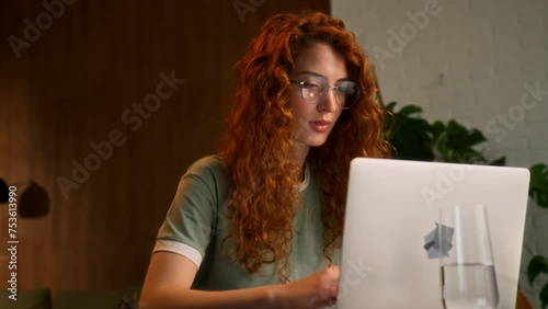 young red haired confident woman sits at table working at laptop at home. creative female student typing on keyboard and looking in screen doing project online education in living room, smiling