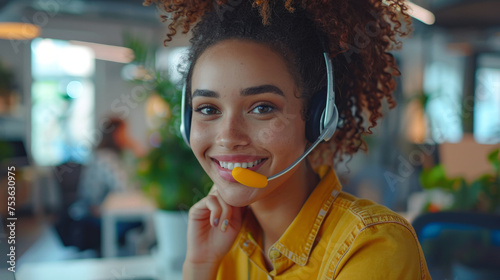 A smiling young woman in a yellow shirt wearing a headset with microphone in a modern office setting.