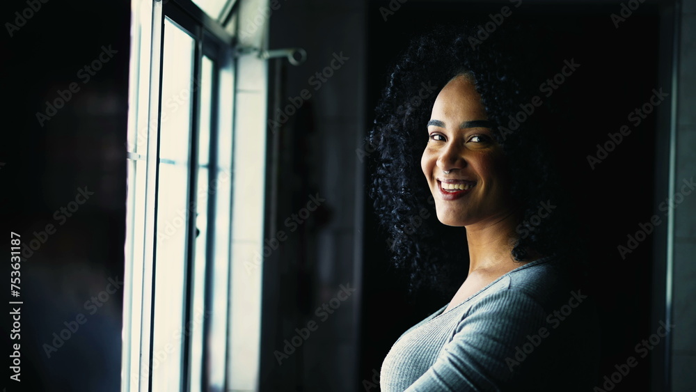 Happy Portrait of a young black Brazilian woman turning head to camera smiling standing by window at home. Curly hair South American adult girl smiling