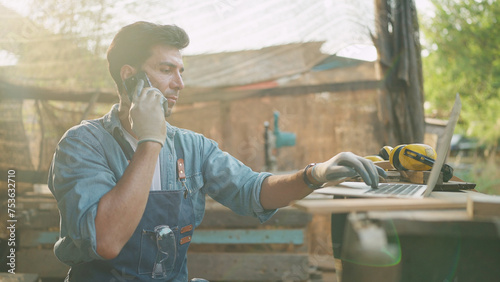 Young carpenter talking on smartphone and using laptop while working on woodworking in the furniture factory. Carpenter and woodcraft concept