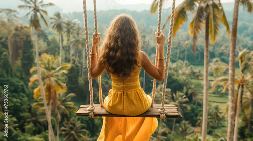Rearview of the young woman with curly brunette hair, wearing a yellow dress and sitting on a wooden swing with ropes, swinging high in the air above the palm trees and meadows on a summer day