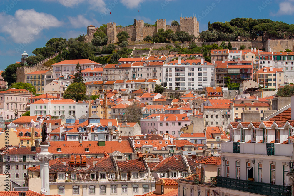 Cityscape of a colorful Lisbon with houses and Castelo de S. Jorge in the background and a blue sky.