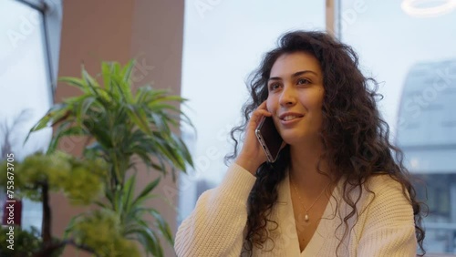 A business woman with brown curly hair seals a deal on the phone, feeling excited, making a winning gesture, dressed in a white sweater with plants and windows in the background photo