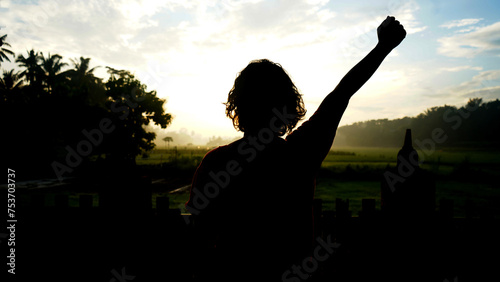 Silhouette of a woman standing alone against the sunrise with one clenched hand raised upwards. Fresh start and ready for new day. Positive self motivation concept background.
