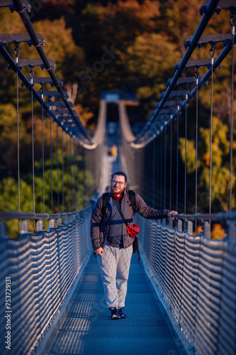 An equipped photographer with a backpack stands ready on a suspension bridge, the golden foliage of autumn setting a vibrant backdrop for adventure © Иванна Емельянова