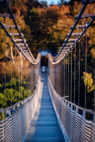 The warm, late-afternoon sun casts a golden glow over an empty suspension bridge, framed by the fiery colors of fall foliage, inviting a tranquil walk © Иванна Емельянова