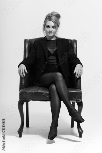 Beauty and fashion concept. Portrait of beautiful woman with tight black clothes and suit sitting on vintage armchair and looking at camera with seductive look. Black and white image