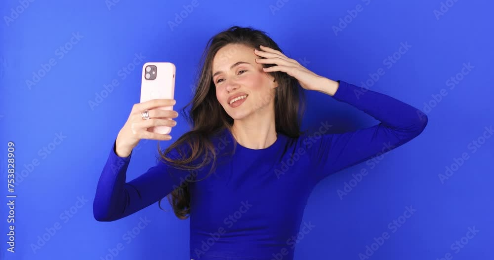 Brunette girl stares at the phone and fixes her hair. Female uses phone like mirror, peace gesture. Young girl make photo from hands with phone on blue background, look at camera and smiling. Brunette girl stares at the phone and fixes her hair. Female uses phone like mirror, peace gesture. Young girl make photo from hands with phone on blue background, look at camera and smiling.