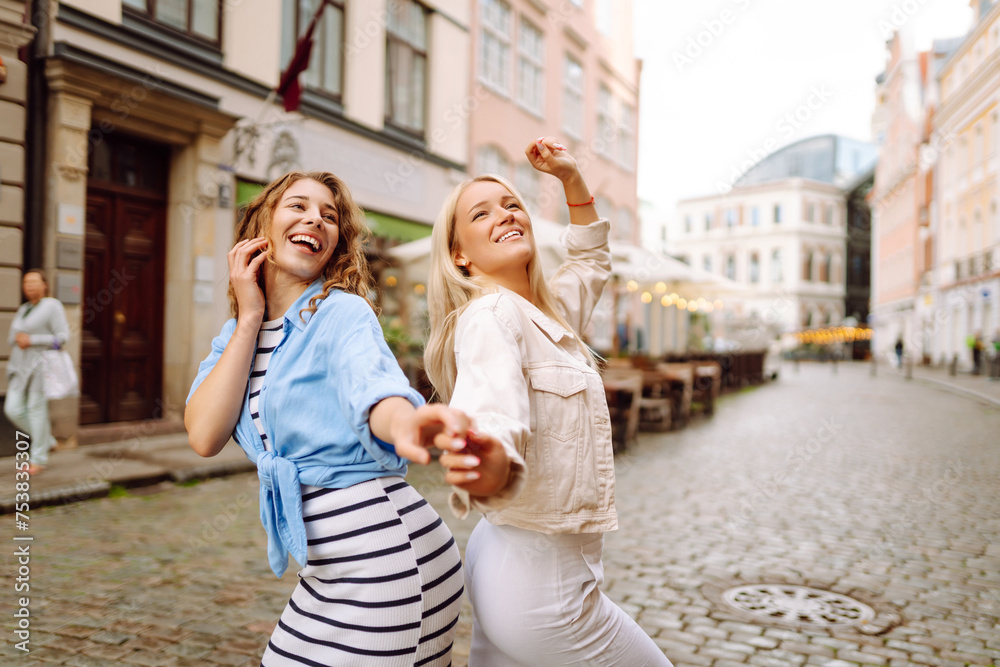 Two young women walk along the city streets. Female tourists in bright dress enjoys sights of the city on sunny day. Concept of lifestyle, fashion, travel.