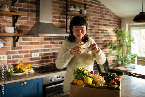 Content Asian woman photographing vegetables in sunlit kitchen © Vorda Berge