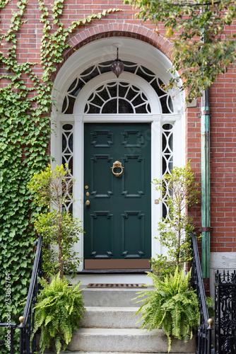 Nestled within a lush embrace, a charming green front door beckons from its set-back position, framed by vibrant foliage and flanked by climbing greenery adorning the classic colonial brick façade. 