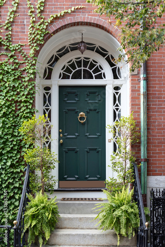 Nestled within a lush embrace, a charming green front door beckons from its set-back position, framed by vibrant foliage and flanked by climbing greenery adorning the classic colonial brick façade.

