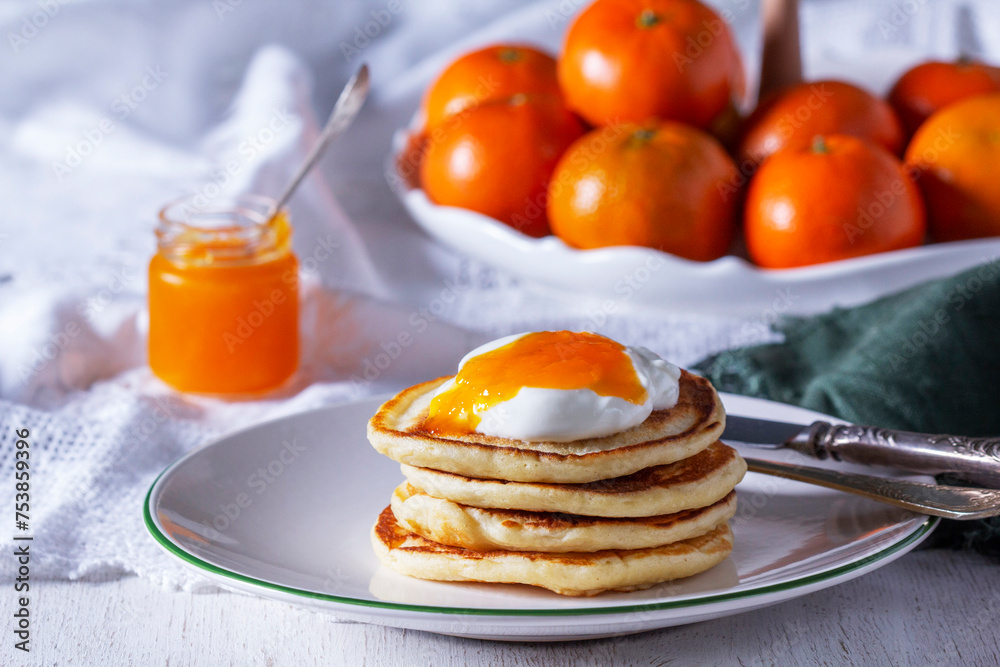 Pancakes served with tangerine jam and sour cream, and tangerines on a light background.