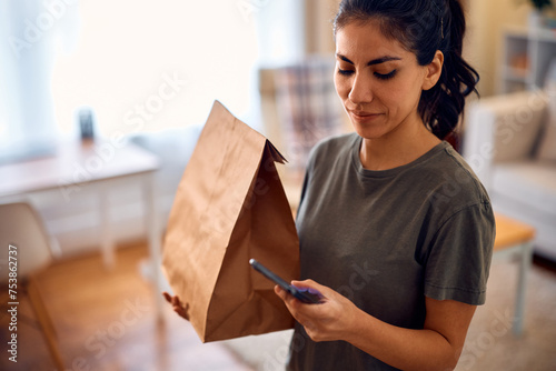 Hispanic woman texting on cell phone after receiving home delivery in paper bag.