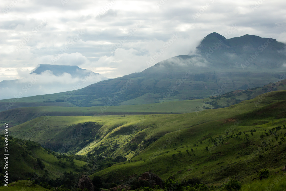 Green sunlit valley, with the mountains in the background shrouded in mist in a early morning in Royal Natal National Park, in the Drakensberg Mountains of South Africa Green sunlit valley, with the mountains in the background shrouded in mist in a early morning in Royal Natal National Park, in the Drakensberg Mountains of South Africa
