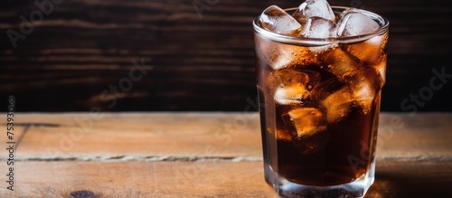 A top-down view of a glass filled with cola and ice cubes placed on a rustic wooden table. The focus is on the clear space around the glass.