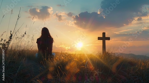 Silhouette Of A Woman Sitting On The Grass Praying In Front Of A Cross At Sunset #753938547