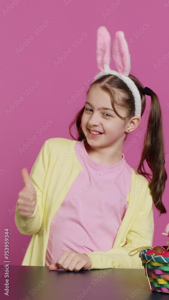 Vertical Video Happy schoolgirl with bunny ears showing thumbs up sign, feeling excited about easter holiday preparation. Cheerful child doing like okay sign and decorating baskets for festivity