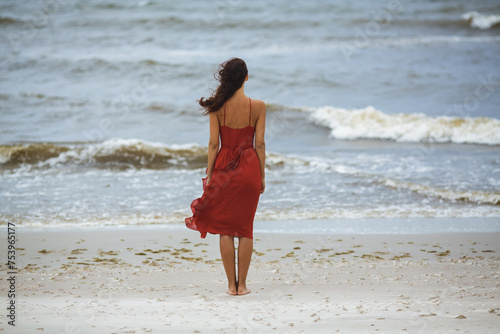 Thoughtful beach woman in red dress standing in cold windy weather. Stormy waves.