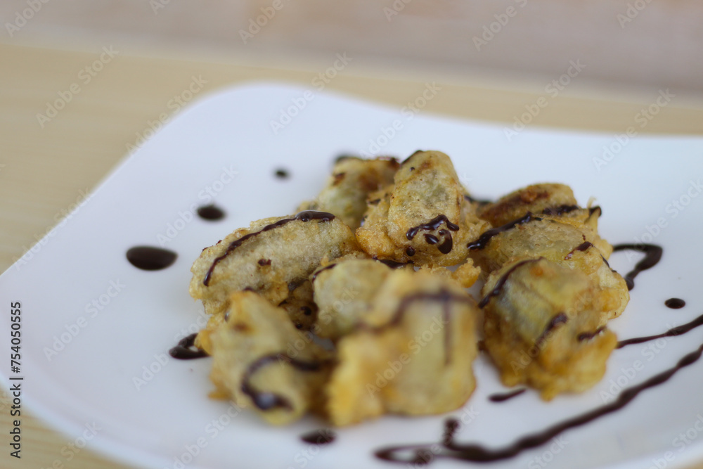 Pisang Goreng Keju or Fried Banana topped chocolate with on white plate on the wooden table. Pisang Goreng Keju or Fried Banana topped chocolate with on white plate on the wooden table.
