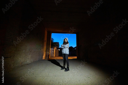 A girl in gray clothes is standing in a doorway. An abandoned industrial building. The time of day is twilight.