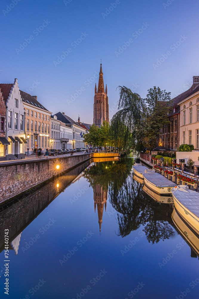 Reflections on a canal in Bruges in the evening. Historic half-timbered houses and the Church of Our Lady in the center of the old town of the Belgian Hanseatic city. Boats and trees with footpath