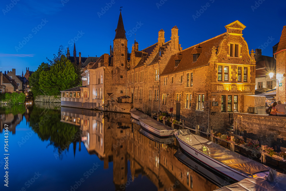 Evening atmosphere in Bruges at the Rosary Quay canal. Illuminated historic buildings of the old town. Reflections of old merchant houses on water surface. Boats at the jetty