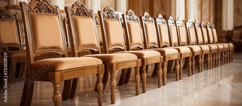 A row of luxurious wooden chairs with beige upholstery lined up next to each other in an assembly hall. The chairs are neatly arranged in a uniform manner, creating a sense of order and sophistication
