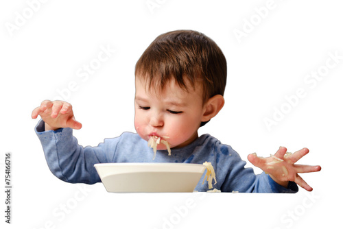 A funny child is eating a grated apple with mouth full while sitting on a kitchen chair, isolated on white background. Hungry baby boy shoves food in his mouth, humor. Kid aged one year four months photo