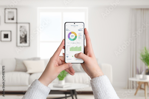 Woman holds smartphone with electricity usage app, monitoring and analyzing consumption for smart home. Living room background. Concept of energy efficiency photo