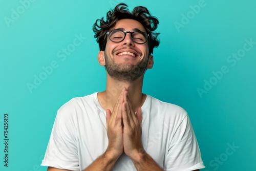A young man with glasses wearing a white t-shirt stands against an isolated background, expressing relief and gratitude with hands on chest