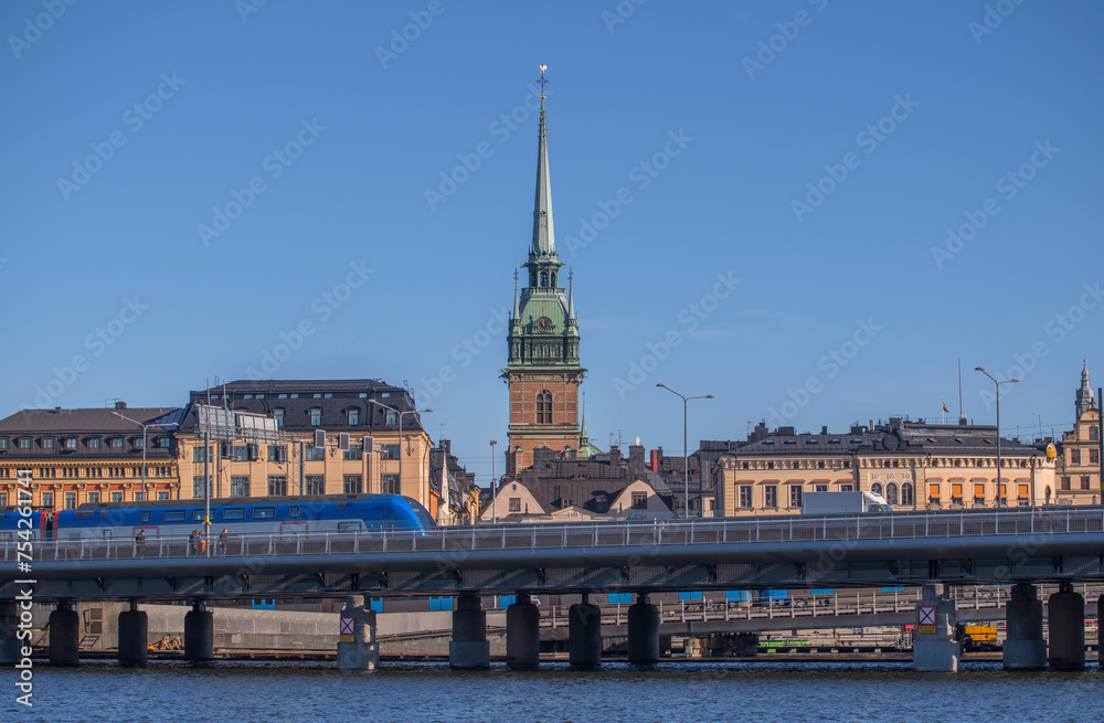Blue commuter train on a bridge passing the old town Gamla Stan and the church tower of Tyska kyrkan, a sunny winter day in Stockholm