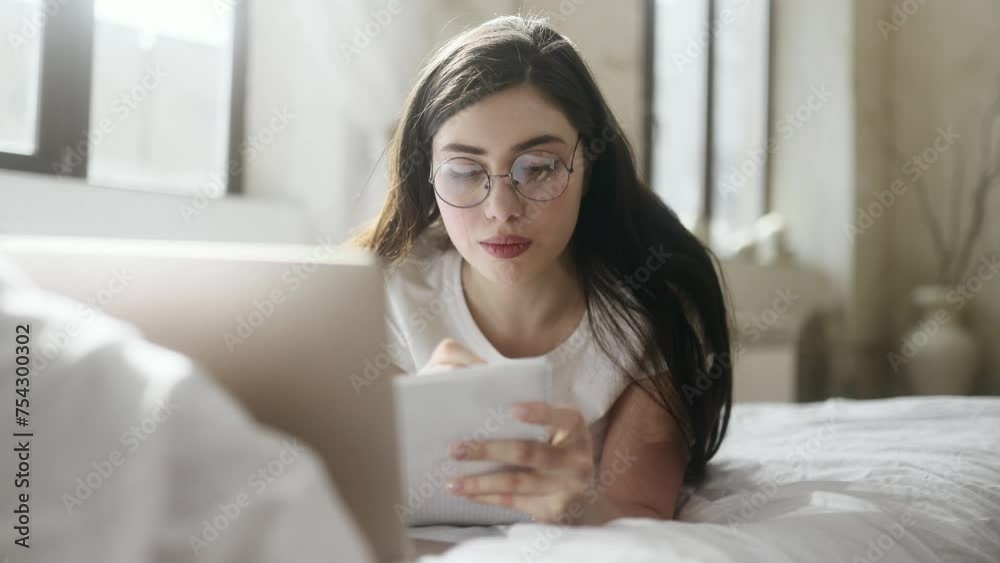 Pretty focused young woman with glasses preparing for exam at university studying laying on bed at home Busy student make notes while looking at laptop computer indoors Distance remote education Pretty focused young woman with glasses preparing for exam at university studying laying on bed at home Busy student make notes while looking at laptop computer indoors Distance remote education