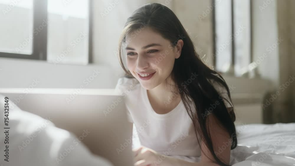 Smiling young brunette woman relaxing at home or working online browsing products in internet store reading news checking email or social media profile on laptop computer laying on bed indoors Smiling young brunette woman relaxing at home or working online browsing products in internet store reading news checking email or social media profile on laptop computer laying on bed indoors