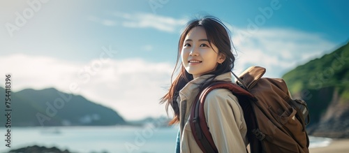 A young Asian woman, wearing a backpack, walks along the sandy beach next to the sea with mountains in the background. The woman is enjoying a leisurely hike as she explores the coastal landscape. © TheWaterMeloonProjec