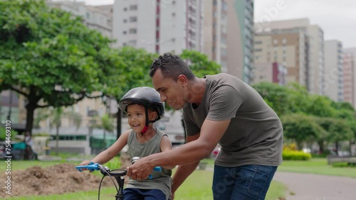 Happy latin father helping smiling boy to ride bike on cityu park. Happy latin child and young dad riding bike. Smiling daddy teaching son to ride a balance bicycle.
